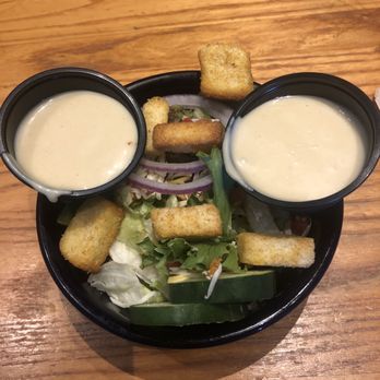 Chili's - Side salad - part of 3 for 10.99 - A bowl of salad containing lettuce, cucumber slices, and red onion rings, topped with several croutons. Two small black cups of creamy dressing are placed on top of the salad, positioned on a wooden table surface. The image captures a typical side salad presentation at a restaurant. - Tex-Mex Near Me - Manteca, California