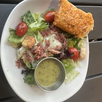 Harveys - Side salad - A plate features a mixed green salad with cherry tomatoes, croutons, and bacon bits. A piece of fried fish is placed on top of the salad, and a small metal cup containing green dressing is positioned on the plate. The dish is served on a white oval plate on a dark wooden surface. - Seafood Near Me - Columbus, Mississippi