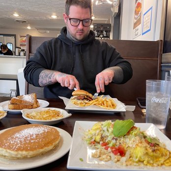 D’ Lua Pancake & Burrito House - taco omelet with pancakes & hashbrowns - A man wearing glasses and a black hoodie is cutting into a burger with fries on a restaurant table, surrounded by breakfast dishes and drinks. - Mexican Near Me - Moline, Illinois