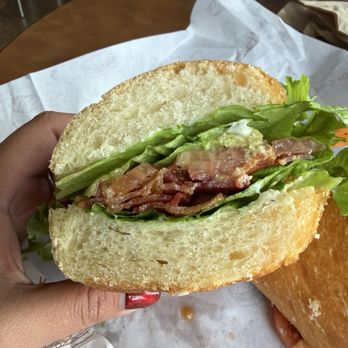 PureBread Deli - A close-up of a sandwich featuring crispy bacon, fresh lettuce, and sliced avocado inside a toasted bun. The sandwich is held by a person with red nail polish, and the background shows a wooden table with paper wrapping and other food items. The setting appears to be a casual dining environment. - Delis Near Me - Newark, Delaware