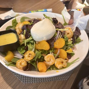 The Kitchen - A seafood salad with cooked shrimp, mandarin orange segments, mixed greens, and a large burrata cheese ball on top. A small container of yellow dressing is placed on the side. The dish is served in a white bowl on a wooden table, with some napkins and utensils nearby. - Bars Near Me - Carmel, Indiana