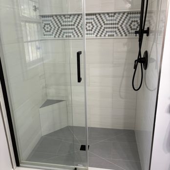 Galenski Tile & Flooring - Shower with stand out Niche - The image shows a walk-in shower with a clear glass door and black hardware, including a showerhead and handle. The shower walls are tiled with large, light-colored tiles, and a horizontal decorative tile strip with geometric patterns runs across the middle. There is a small built-in corner seat in the shower. The floor is tiled with gray tiles, and a black drain is visible. A window with a grid pattern is reflected in the glass, indicating natural light in the space. - Tiling Near Me