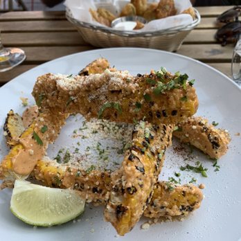 FancyFree - Elote ribs - A white plate holds elote ribs coated with cheese, herbs, and seasoning, garnished with a lime wedge. The ribs are arranged in a circular pattern on the plate. In the background, a basket of fried food with dipping sauce is visible, placed on a wooden table. The setting appears to be an outdoor dining area. - Pubs Near Me - Brooklyn, New York