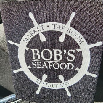 Bob's Seafood - Koozies for the win! - A fabric cover displays a boat wheel logo with the text 'Bob's Seafood' in the center. Surrounding the logo are the words 'Market,' 'Tap Room,' and 'Restaurant,' indicating different areas or services of the seafood business. The cover appears to be placed on a surface, possibly in a vehicle or outdoor setting, with textured background material visible. - Seafood Markets Near Me - Windham, Maine