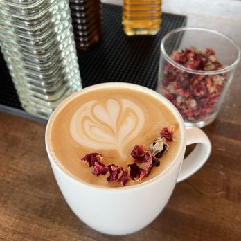 Third Way Cafe - Golden Rose Latte - A latte with foam art in a white ceramic mug, decorated with dried rose petals on top, placed on a wooden table. In the background, there are glass jars, bottles, and a container filled with dried rose petals, indicating a coffee shop setting. - Coffee & Tea Near Me - Columbus, Ohio