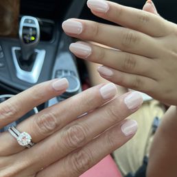 The image features two hands with manicured nails painted in a light, neutral shade. One hand displays a wedding ring with a large central stone surrounded by smaller stones. The background shows the interior of a car, including part of the gear shift and dashboard. The focus is on the hands and nails, with the car interior slightly blurred.