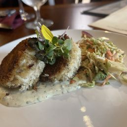 Crab Cakes - Two crab cakes garnished with microgreens are placed on a white plate, accompanied by a side of coleslaw with shredded vegetables and dressing. The crab cakes are breaded and fried, with a creamy sauce underneath. The background shows a wooden table, wine glasses, and menus, indicating a restaurant environment.