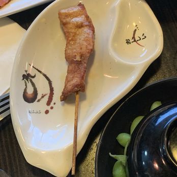 Osaka - A piece of grilled meat on a wooden skewer is placed on a Japanese-style ceramic plate with Japanese characters and artistic design. The plate is part of a meal setting that includes other dishes, such as a bowl of edamame and a bowl of soup, indicating a Japanese cuisine dining experience. - Asian Fusion Near Me - Bloomfield, Connecticut