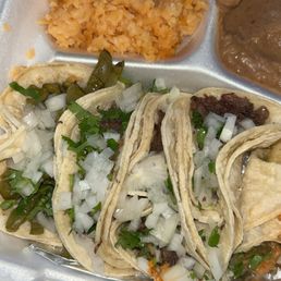 5 taco plate $13.99 - The image shows a styrofoam tray with three tacos filled with meat, chopped onions, and cilantro. The tacos are arranged side by side, with some green peppers visible. In the upper section of the tray, there is a portion of rice and a serving of refried beans. The tray appears to be a takeout meal from a Mexican restaurant or taco spot.