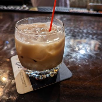Celtic Crossing Irish Pub - A clear glass filled with iced coffee, ice cubes, and a red straw is placed on a coaster on a dark, polished wooden bar counter. The background shows a blurred figure and bar area, indicating a casual dining or drinking environment. - Irish Pub Near Me - Chicago, Illinois