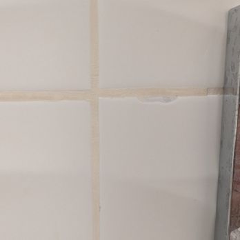 Jesse & Bro’s Construction - A close-up image of a tiled wall featuring white ceramic tiles with beige grout lines. One tile has a small chip or damage along its edge, indicating minor wear or defect. The tiles are arranged in a grid pattern, and part of a metal object is visible on the right side of the image. - General Contractors Near Me - Oxnard, California