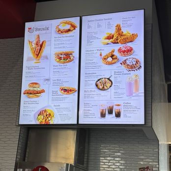 Bruxie - Menu - A large digital menu screen at Bruxie restaurant in Davis, CA, presents a variety of food items such as fried chicken sandwiches, waffle sandwiches, chicken tenders, waffles, and sides. The menu includes images, item descriptions, and prices, with sections for different meal options and beverages. The display is mounted on a wall with a tiled background and a Coca-Cola beverage dispenser below it. - Chicken Shop Near Me - Davis, California