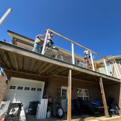 Nick's Construction And Remodeling - Three workers on ladders are constructing a roof extension or porch cover on a brick house under a clear blue sky...