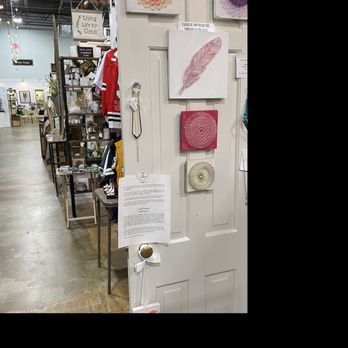 Painted Tree Kildeer - A display of textured wall art pieces is arranged on a white door in a retail store. The artwork includes circular designs in pink, purple, and cream, and a feather-shaped piece in pink. The store interior features shelves with various items, clothing, and signs, with a visible industrial ceiling and lighting. A printed notice is attached to the door handle, and the display appears to be part of an art or home decor section. - Home Decor Near Me - Kildeer, Illinois