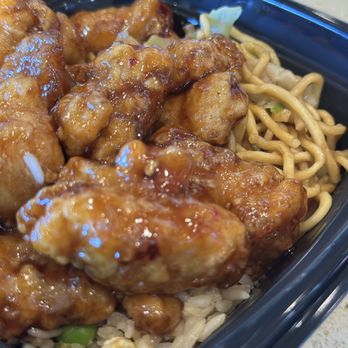 Panda Express - Orange Chicken Bowl w/ Fried Rice & Chow Mein - The image shows a black plastic takeout container filled with Asian food. The dish includes orange chicken coated in a shiny, sticky sauce, fried rice with small pieces of vegetables, and chow mein noodles. The food is arranged with the chicken on top, partially covering the rice and noodles. The container is placed on a light-colored surface, and the focus is on the food's textures and colors. - Chinese Near Me - Sonora, California