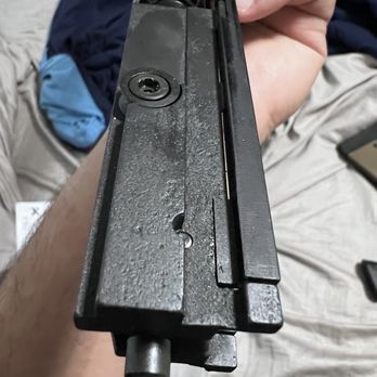 Utas-Usa - The image depicts a close-up view of a firearm's slide and bolt assembly, focusing on the top side. The metal surface shows machining marks and a textured finish. The slide features a visible ejection port and a rear sight. The background includes a person's hand holding the firearm, with part of their arm and casual clothing visible. The setting appears to be indoors, possibly in a home environment. - Guns & Ammo Near Me - Des Plaines, Illinois