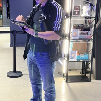 Sandbox VR - This is the rude person/manager I was referring to. - A young man with short dark hair is standing indoors, wearing a black jacket with white stripes on the sleeves, a black t-shirt, and blue jeans. He is holding a tablet device and appears to be focused on it. Behind him, there is a display shelf with various items, including a mannequin head with a cap, boxes, and other merchandise. The setting appears to be a retail or entertainment venue with modern lighting and decor. - Virtual Reality Centers Near Me - Chicago, Illinois