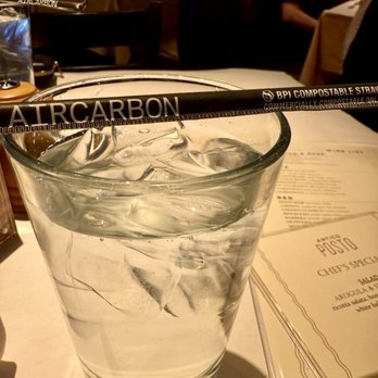 Antico Posto - I found it interesting that they use BPI compostable straws - A tall, transparent glass contains water with several ice cubes, positioned on a restaurant table. A black BPI compostable straw is laid across the top of the glass. The background shows a dimly lit restaurant interior with tables, chairs, and a person seated at a table. The setting appears to be a dining area with ambient lighting and a menu partially visible on the table. - Italian Near Me - Oak Brook, Illinois
