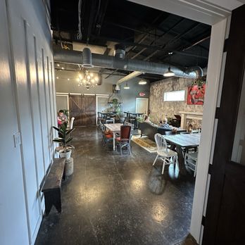 Grutogi Bistro - The image shows an indoor dining space with a polished dark floor, featuring various tables and chairs, including a white wooden chair and a cushioned armchair. The ceiling has exposed black ductwork and industrial-style lighting, with a chandelier hanging in the center. The walls include a textured stone finish and a colorful abstract painting. Potted plants are placed along the wall, and a sliding barn door is visible at the back, suggesting a cozy, eclectic atmosphere. - Coffee & Tea Near Me - Keller, Texas