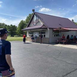 Outside area - A small ice cream stand named 'Twist' is situated in an outdoor area on a sunny day with a partly cloudy sky. Several customers are standing in line or ordering at the window, while others are seated at picnic tables under a striped canopy. The stand has a gabled roof and is surrounded by a paved parking area and green trees in the background.