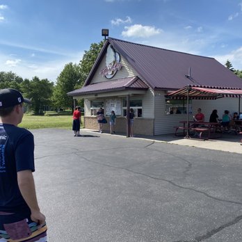 Mr. Twisty - Outside area - A small ice cream stand named 'Twist' is situated in an outdoor area on a sunny day with a partly cloudy sky. Several customers are standing in line or ordering at the window, while others are seated at picnic tables under a striped canopy. The stand has a gabled roof and is surrounded by a paved parking area and green trees in the background. - Ice Cream & Frozen Yogurt Near Me - St  Cloud, Minnesota