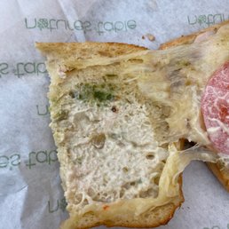 A close-up of a sandwich slice with melted cheese, chopped vegetables, and a tomato slice on top, placed on branded paper.