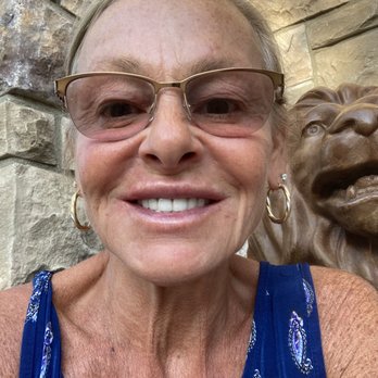 Evolve Med Spa - A woman wearing glasses and hoop earrings is smiling at the camera, with a stone wall and lion sculpture in the background. - Laser Hair Removal Near Me - Hoboken, New Jersey