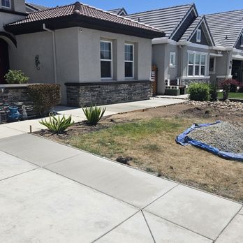 HSD Masonry - The image shows the front yard of a residential house with a concrete sidewalk and a small landscaped area with plants. Part of the yard has exposed soil and a blue tarp, suggesting ongoing landscaping or yard work. The house has a modern exterior with a stone accent wall and multiple windows, under a clear blue sky. - Masonry/Concrete Near Me