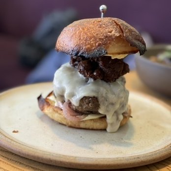 Gator - A burger with a fried chicken patty, melted cheese, and toppings, assembled on a bun with a toothpick securing it, served on a ceramic plate. The burger includes a toasted bun, fried chicken, cheese, and additional toppings, with a side dish visible in the background. - Desserts Near Me - Brooklyn, New York