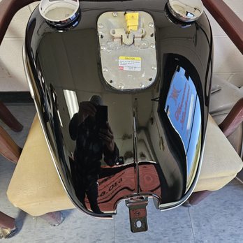 Havana Kustoms - 2006 Softail Springer Classic - A glossy black motorcycle fuel tank with chrome accents and two gauges is placed on a chair. The tank's surface is highly reflective, showing the reflection of the person taking the photo, the ceiling lights, and a nearby sign. The tank has a mounting bracket at the bottom and a yellow safety label on the top. The setting appears to be indoors, possibly in a workshop or showroom. - Body Shops Near Me - Addison, Illinois