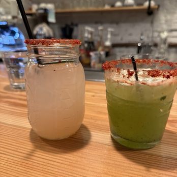 Un Amor - Paloma and Cucumber Margarita - Two drinks are positioned on a wooden table in a restaurant or bar environment. The drink on the left is in a mason jar with a black straw and a salted rim. The drink on the right is in a glass, also with a salted rim, containing a green-colored beverage with ice. The background shows a bar area with shelves, glasses, and various bar equipment, with a slightly blurred focus. - Breakfast & Brunch Near Me - Chicago, Illinois