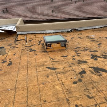 AJ's Professional Contracting - 2-Old roof removed - The photo depicts a rooftop with plywood sheathing, showing signs of recent or ongoing repair work. A skylight is installed on the roof, and a damaged parapet wall with torn material is visible. The background includes neighboring rooftops with red tiles and a mountain range under a cloudy sky. The scene suggests construction or renovation activity on the roof. - Roofing Near Me
