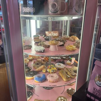 Voodoo Doughnut - Display case - The image shows a glass display case containing an assortment of donuts with various toppings, including sprinkles, icing, and candies, arranged on pink paper. The top shelf holds coffee mugs with the Voodoo Doughnut logo. The display is part of the Voodoo Doughnut shop in Austin, TX, known for creative donut flavors. The case is illuminated, highlighting the colorful and diverse selection of donuts. - Donuts Near Me - Austin, Texas