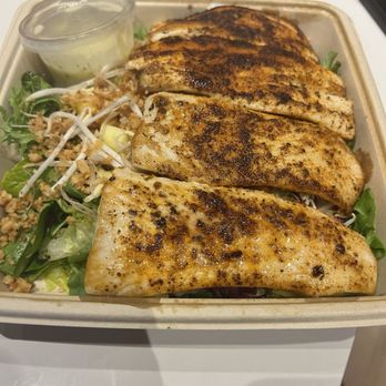Surfside Taco Stand - Tahiti with blackened mahi mahi - The image shows a takeout container with three grilled mahi mahi fillets, each with a seasoned, slightly charred surface. The fillets are placed on a bed of mixed greens and bean sprouts, with a side of creamy dipping sauce in a small plastic cup. The container is on a white surface, and part of a plastic lid and a black object are visible in the background. - Tacos Near Me - Nantucket, Massachusetts