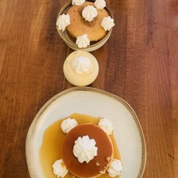 Flan - The image shows three desserts on a wooden table. The largest dessert is a flan with caramel sauce, topped with whipped cream and surrounded by additional whipped cream dollops. A smaller pudding with whipped cream is in a glass container. A cookie with whipped cream dollops on top is served in a small bowl. The desserts are arranged in a vertical line, with a spoon visible at the top of the image.