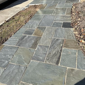 CM Hardscapes - Flagstone patio walkway - A flagstone patio walkway features irregular gray and brown stone tiles, bordered by soil and adjacent to a driveway with a parked car. - Masonry/Concrete Near Me
