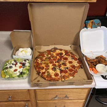 Sergio's Bakery and Cafe - Large pizza with pepperoni, mushroom, black olive, green chile, buffalo wings with ranch, Greek salad, and a lemon creme pie. - A pizza with various toppings including pepperoni, mushrooms, black olives, and green chile is placed in an open cardboard box on a kitchen countertop. To the left, there is a fresh salad with lettuce, cherry tomatoes, black olives, and shredded cheese in a plastic container, along with a small packet of Italian seasoning and a dressing cup. Behind the salad, a dessert with whipped cream and lemon slices is in a small box. To the right of the pizza, a white takeout container holds chicken wings with two small cups of dipping sauce and a couple of baby carrots. The scene is set on a kitchen counter with a stove and a red wall in the background. - Bakeries Near Me - Albuquerque, New Mexico
