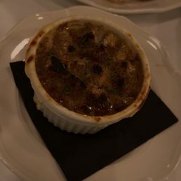 Onion Soup Gratinée - A serving of French onion soup topped with melted cheese, served in a ramekin on a black napkin, with a plate of bread in the background.