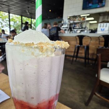 Showroom - Milkshake - Strawberry Marshmallow - September 2025 - A strawberry milkshake with a layer of red strawberry syrup at the bottom, topped with whipped cream and a crumbly garnish, is presented in a tall glass with a green-and-white striped straw. The background shows a modern restaurant interior with large windows, a bar area, and patrons dining, indicating a casual dining environment. - New American Near Me - Frederick, Maryland