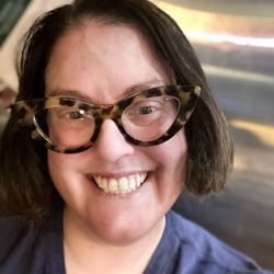 SEE - A white-passing woman with dark brown hair wearing cat-eye tortoiseshell glasses and a blue shirt.