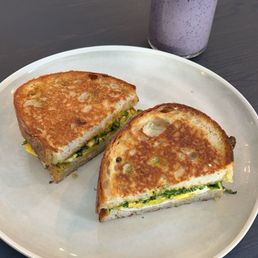 Egg Melt - A plate with two grilled sandwiches filled with vegetables and eggs, accompanied by a purple smoothie topped with yellow sprinkles, on a dark table at a café.