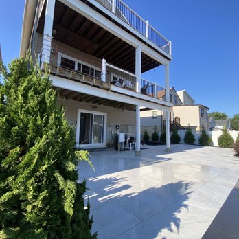 Masone Masonry - The image shows a backyard with a large concrete patio area and a multi-level wooden deck attached to a house. The deck has railings and outdoor furniture, with stairs leading down to the patio. Green bushes and trees border the space, and the sky is clear and blue, indicating a sunny day. The setting appears to be a residential outdoor area designed for leisure and outdoor activities. - Masonry/Concrete Near Me