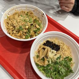 Sichuan Cold Noodles - Two bowls of Asian noodle dishes with toppings like peanuts, herbs, and sauces on a red tray, with a person holding chopsticks above.