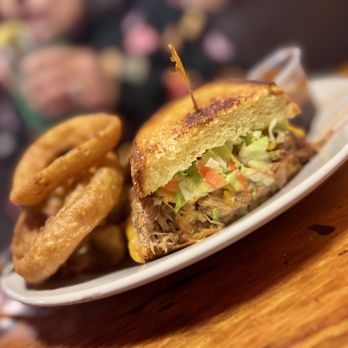 Rainbow Oaks Restaurant - Pork Sandwich with fries & onion rings - A sandwich with shredded pulled pork, lettuce, tomato, and sauce is presented on toasted bread, accompanied by onion rings and fries on a white plate. The plate is placed on a wooden table, with a blurred background indicating a restaurant setting. The image highlights the food items served at Rainbow Oaks Restaurant in Fallbrook, CA. - American Near Me - Fallbrook, California