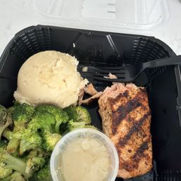 Salmon - A takeout meal in a black plastic container includes mashed potatoes, steamed broccoli, grilled salmon, and a small cup of sauce.