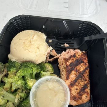 Bonefish Grill - Salmon - A takeout meal in a black plastic container includes mashed potatoes, steamed broccoli, grilled salmon, and a small cup of sauce. - Seafood Near Me - Independence, Ohio