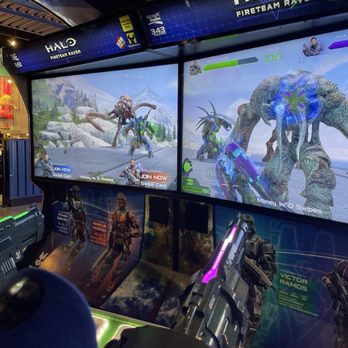Playdium - Two large arcade screens show a sci-fi shooting game with characters and monsters in a snowy landscape. Players hold laser guns, aiming at the targets on the screens. The environment includes gaming stations with character posters and a visible area of the arcade with other patrons and colorful decorations. The setting is an entertainment venue in Whitby, ON, offering interactive gaming experiences. - Arcades Near Me - Whitby, Ontario