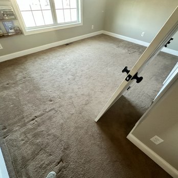 Precision Property Service - The image shows an empty room with beige carpet flooring that has visible indentations and marks. There is a large window on one wall, allowing natural light to illuminate the space. A partially open door is visible, with a backpack hanging on the door handle. The walls are painted a neutral color, and there are electrical outlets and a vent on the floor. The room appears clean and ready for furnishing or remodeling. - Drywall Installation & Repair Near Me - Plainfield, Connecticut