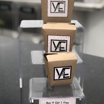 Y&E Creations - three boxes stacked on top of each other - Three small cardboard boxes with VE Creations labels are stacked vertically on a clear acrylic display stand. The labels feature a black and white logo with the letters 'VE' and an ampersand. A promotional card below the boxes advertises a buy 9, get 1 free offer, with empty circles for stamps or signatures. The background appears to be a kitchen or food preparation area, with blurred metallic surfaces and equipment. - Desserts Near Me - Middletown, New York