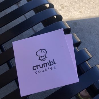 Crumbl - Naperville - A white box featuring the Crumbl Cookies logo, including a cartoon cookie character, is positioned on a black metal bench outdoors. The bench has horizontal slats, and the background shows concrete pavement with sunlight casting shadows. - Bakeries Near Me - Naperville, Illinois